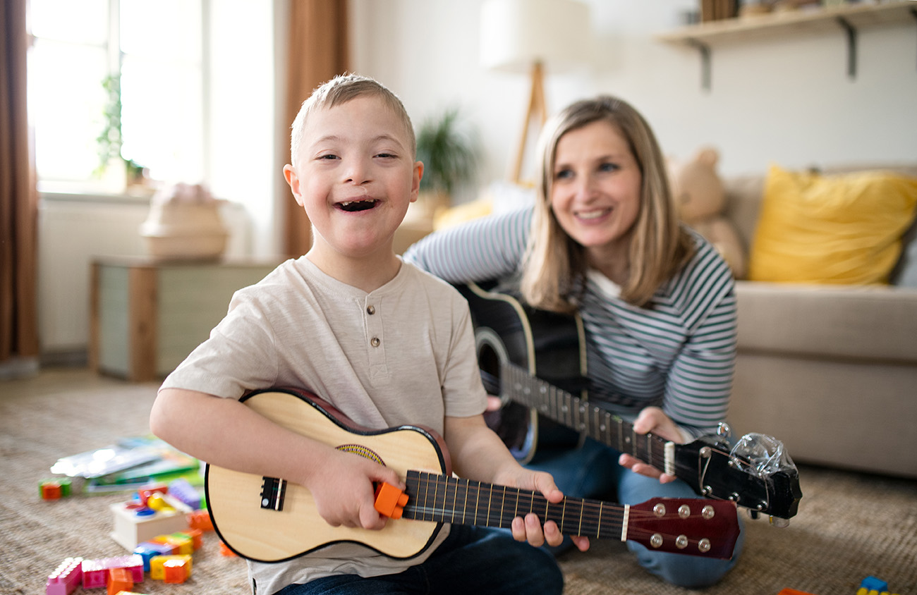 Fröhliches Kind mit Down-Syndrom sitzt auf dem Boden und spielt mit Begeisterung eine kleine Gitarre (Ukulele). Im Hintergrund sitzt eine lächelnde Frau, vermutlich die Mutter, die ebenfalls eine Gitarre hält. Um sie herum liegen bunte Spielsteine, was auf eine gemütliche und spielerische Atmosphäre im Wohnzimmer hinweist.