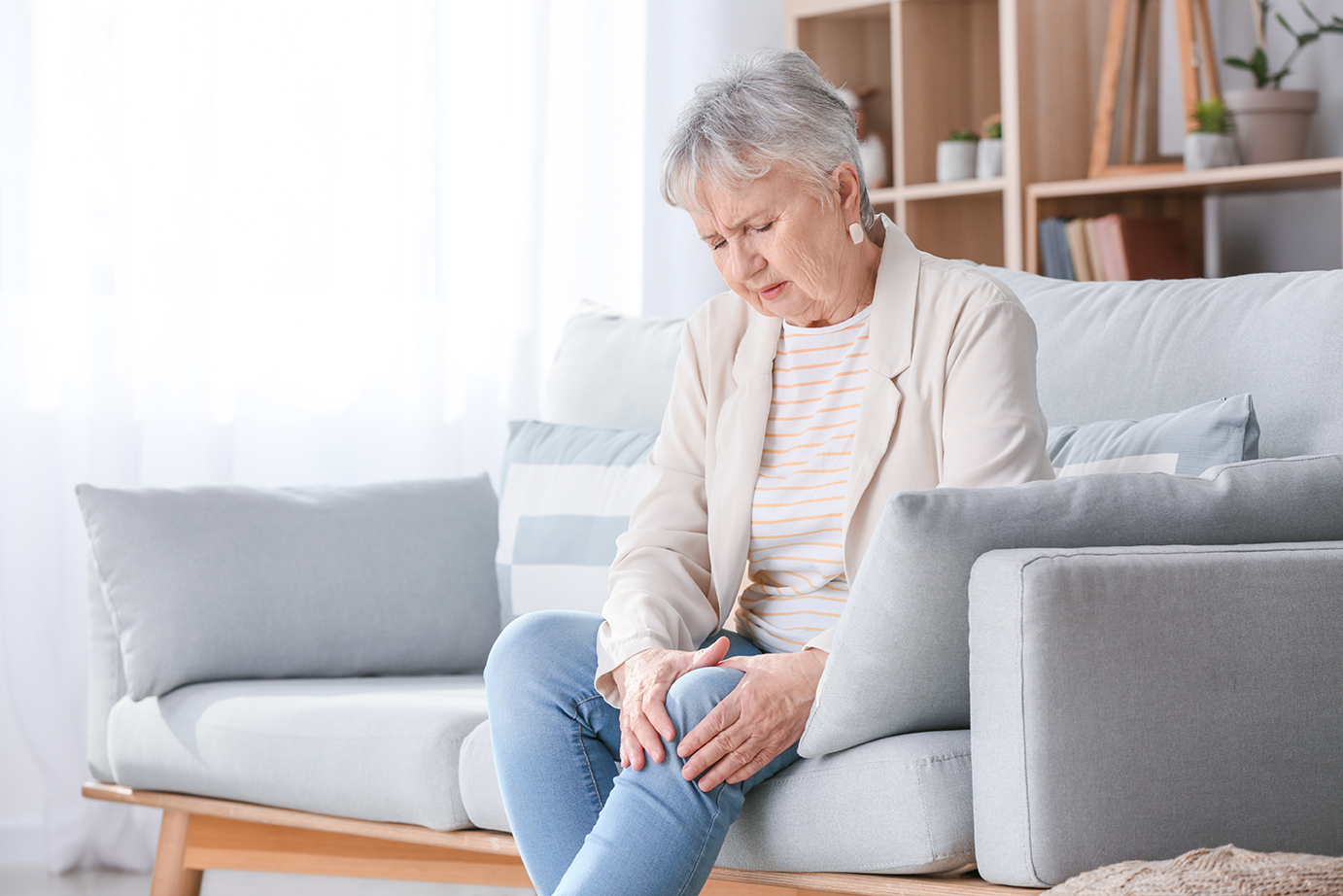 Eine ältere Frau sitzt auf einem Sofa in einem gemütlich eingerichteten Wohnzimmer. Sie hält sich mit schmerzverzerrtem Gesicht das Knie und scheint unter Gelenkbeschwerden oder Arthritis zu leiden. Im Hintergrund sind ein Regal mit Büchern und Pflanzen sowie weiche Kissen auf dem Sofa zu sehen.