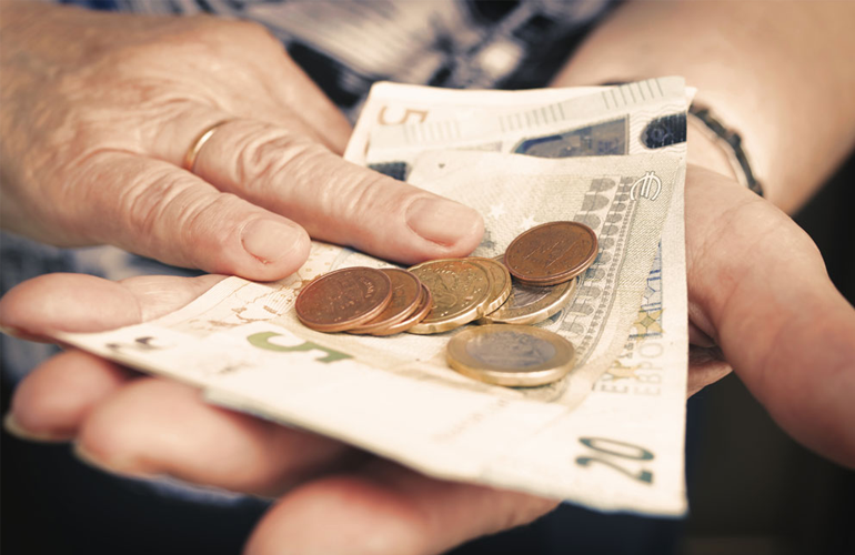 Nahaufnahme von älteren Händen, die einige Euro-Scheine (5 und 20 Euro) sowie verschiedene Euro-Münzen halten. Eine Hand stützt die Scheine, während die andere Hand leicht darüber liegt. Die Person trägt einen schlichten Ring am Finger.