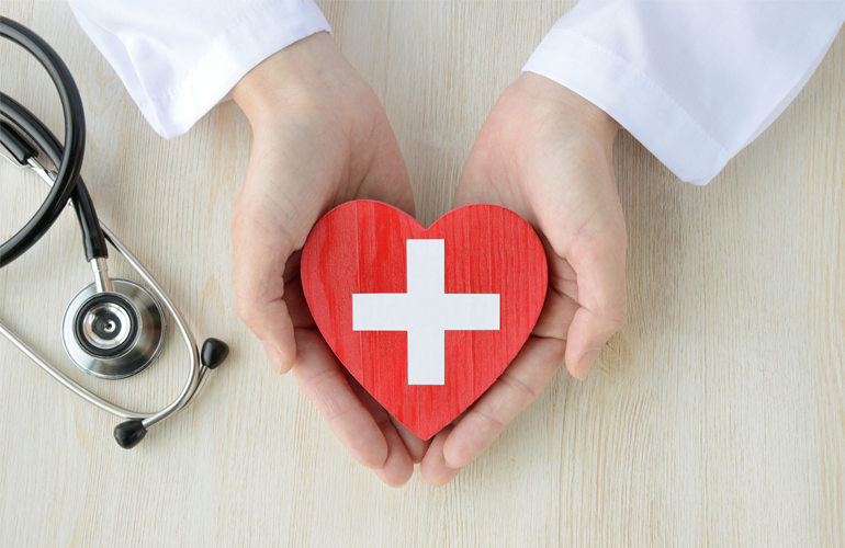 Hände in einem weißen Arztkittel halten ein rotes, herzförmiges Symbol mit einem weißen Kreuz in der Mitte, das für Gesundheit und medizinische Versorgung steht. Links im Bild liegt ein Stethoskop auf einer hellen Holzoberfläche.