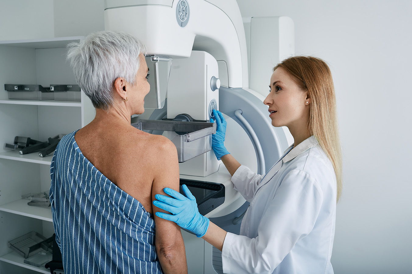 Eine ältere Frau mit kurzen grauen Haaren steht vor einem Mammographie-Gerät zur Brustkrebsfrüherkennung. Sie trägt ein gestreiftes medizinisches Oberteil, das eine Schulter freilässt. Eine Ärztin oder medizinische Fachkraft mit langen blonden Haaren und blauen Handschuhen erklärt den Ablauf der Untersuchung und justiert das Gerät. Die Szene findet in einer hellen, modernen medizinischen Umgebung statt.