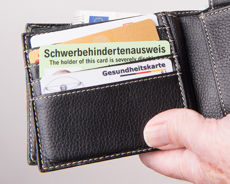Nahaufnahme einer schwarzen Leder-Geldbörse, die mehrere Kartenfächer enthält. Sichtbar sind ein Schwerbehindertenausweis in hellgrün mit der Aufschrift ‚The holder of this card is severely disabled‘, eine Gesundheitskarte sowie weitere Karten und Dokumente. Eine Hand hält die Geldbörse.