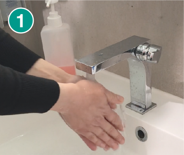 Nahaufnahme von zwei Händen, die unter fließendem Wasser an einem modernen Waschbecken mit einem silbernen Wasserhahn gewaschen werden. Im Hintergrund steht eine transparente Seifenflasche mit rosa Flüssigseife. Die Szene ist mit einer „1“ in einem grünen Kreis markiert, was den ersten Schritt einer Händewasch-Anleitung symbolisiert.