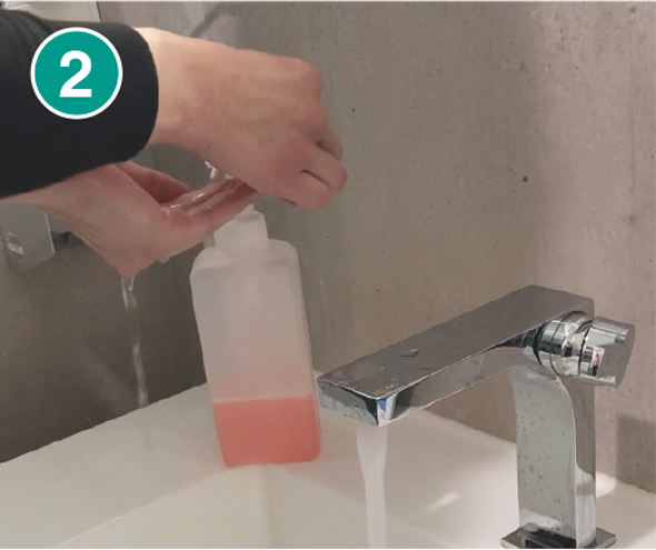 Nahaufnahme von zwei Händen, die Seife aus einem transparenten Seifenspender mit rosa Flüssigseife entnehmen. Der Seifenspender steht auf einem weißen Waschbecken, aus dessen modernem, silbernen Wasserhahn Wasser fließt. In der linken oberen Ecke befindet sich eine grüne Markierung mit der Zahl „2“, die den zweiten Schritt einer Händewasch-Anleitung kennzeichnet.