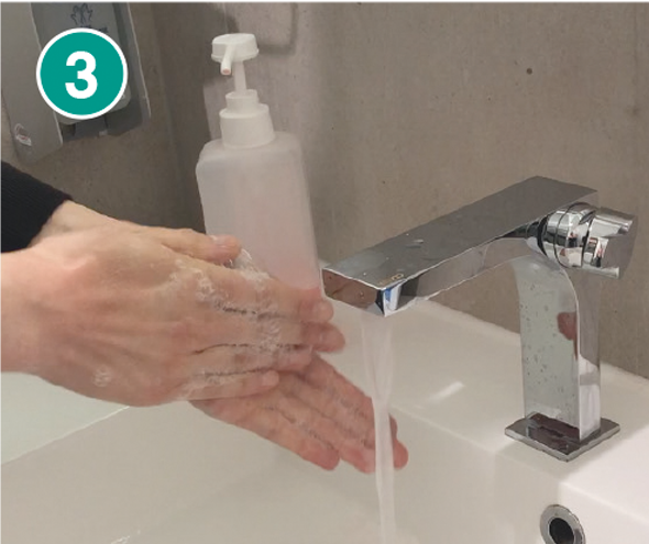 Nahaufnahme von zwei Händen, die mit Seife eingeschäumt werden. Im Hintergrund steht ein transparenter Seifenspender mit weißem Pumpkopf auf einem weißen Waschbecken. Aus dem modernen, silbernen Wasserhahn fließt Wasser. In der linken oberen Ecke befindet sich eine grüne Markierung mit der Zahl „3“, die den dritten Schritt einer Händewasch-Anleitung kennzeichnet.