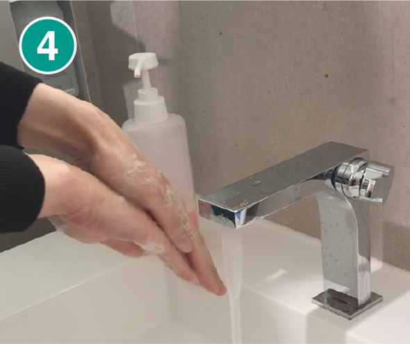 Nahaufnahme von zwei Händen, die gründlich mit Seife eingerieben werden. Die Hände befinden sich über einem weißen Waschbecken mit einem modernen, silbernen Wasserhahn, aus dem Wasser fließt. Im Hintergrund steht ein transparenter Seifenspender mit weißem Pumpkopf. In der linken oberen Ecke des Bildes ist eine grüne Markierung mit der Zahl „4“ zu sehen, die den vierten Schritt einer Händewasch-Anleitung kennzeichnet.