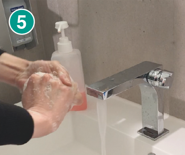 Nahaufnahme von zwei Händen, die gründlich mit Seife eingerieben werden. Die Hände sind zu einer Reibbewegung verschränkt und bilden dabei Schaum. Im Hintergrund ist ein weißes Waschbecken mit einem modernen, silbernen Wasserhahn zu sehen, aus dem Wasser fließt. Ein transparenter Seifenspender mit rosa Seife und weißem Pumpkopf steht am Beckenrand. In der linken oberen Ecke des Bildes befindet sich eine grüne Markierung mit der Zahl „5“, die den fünften Schritt einer Händewasch-Anleitung kennzeichnet.
