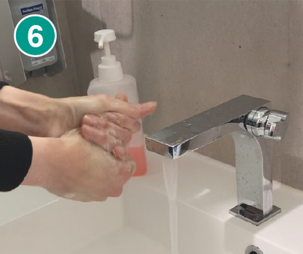 Nahaufnahme von zwei Händen, die gründlich gewaschen werden. Die Hände sind ineinander verschränkt und mit Seifenschaum bedeckt, während sie unter fließendem Wasser von einem modernen, silbernen Wasserhahn abgespült werden. Im Hintergrund steht ein transparenter Seifenspender mit rosa Flüssigseife und einem weißen Pumpkopf. In der linken oberen Ecke des Bildes befindet sich ein grüner Kreis mit der weißen Zahl „6“, der den sechsten Schritt einer Händewasch-Anleitung markiert.