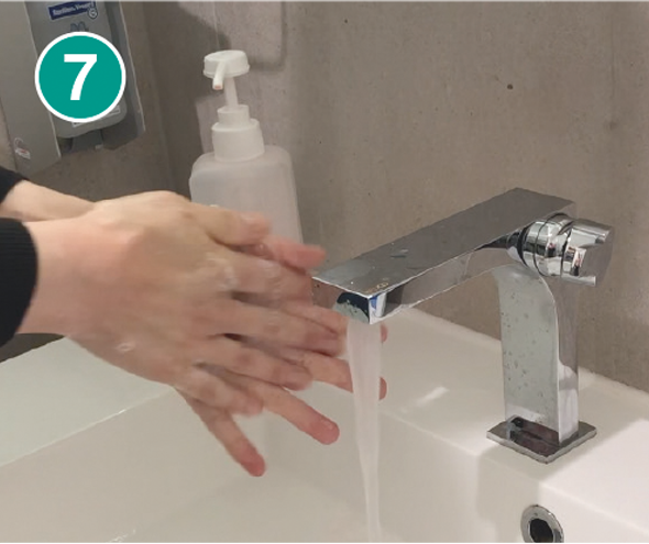 Nahaufnahme von zwei Händen, die unter fließendem Wasser von einem modernen silbernen Wasserhahn abgespült werden. Die Hände reiben sich gegenseitig, um Seifenreste zu entfernen. Im Hintergrund steht ein transparenter Seifenspender mit einem weißen Pumpkopf. In der linken oberen Ecke des Bildes befindet sich ein grüner Kreis mit der weißen Zahl „7“, der den siebten Schritt einer Händewasch-Anleitung markiert.